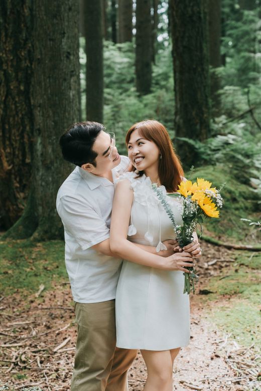 Richard hugging Mabel from behind in a tall pine forest, Mabel holding sunflowers